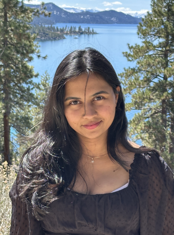 Person in black top with long dark hair smiling outdoors in bright sunlight, with a lake, pine trees, and snow-capped mountains in the background.