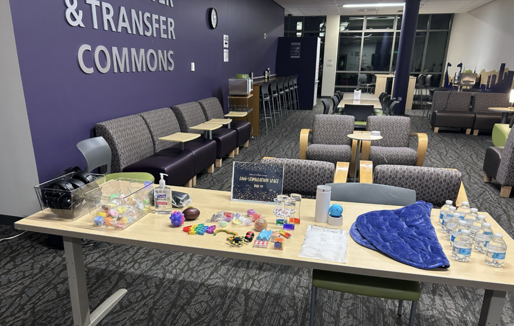 A table displays various items including sensory toys, hand sanitizer, water bottles, and a blue blanket, set against a backdrop of purple walls with "& TRANSFER COMMONS" text and seating areas.
